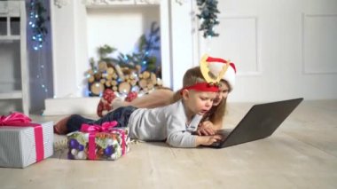 Mutlu küçük çocuklar Noel arifesinde şöminenin yanındaki ağacın altında dizüstü bilgisayar izliyorlar. Geleneksel şömineli dekore edilmiş oturma odası. Evde sıcak bir kış akşamı..