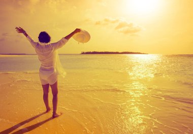 Beyaz gömlekli, kollarını kaldırıp tropik bir sahilde şapkasını tutan ve ufka bakan beyaz bir kadın.