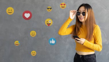 Cep telefonu tutan genç bir kadın, emoji ikonlarına bakıyor. Gri arka planda izole edilmiş, simgeleri olan Asyalı bir kız.