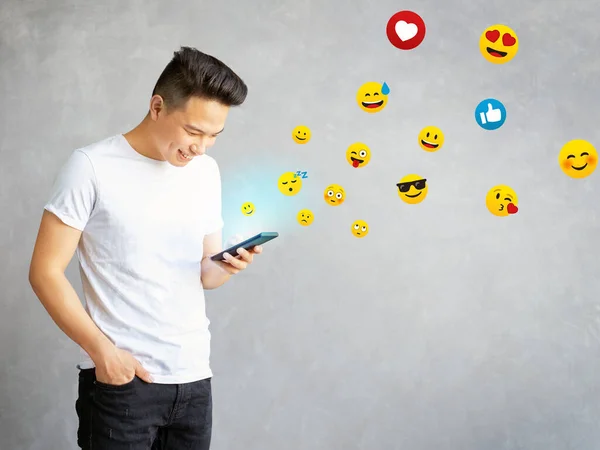 asian guy typing emoji on cell phone, young man having fun using smart phone isolated on grey background