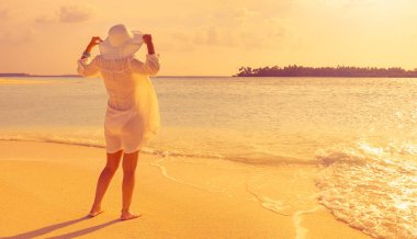 Şık bir kız, beyaz şapkası ve gömleği yumuşak kumda olan bir yaban ördeği adasının tropikal plajında gün batımını izler.