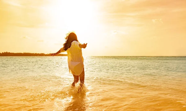 Kız gün batımında beyaz bir bikini ve saçları rüzgarda uçuşan gömleğiyle bir maldivlerin berrak sularında koşuyor.