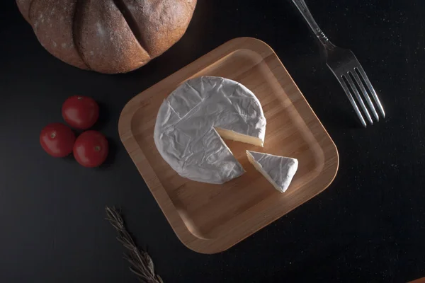 Queso Brie francés fresco y una rebanada en una tabla de bambú con tomates cherry, pan en el plato negro. Puesta plana. — Foto de Stock