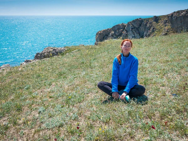 Mature woman, wearing blue fliss jacket, sits on green grass. Azure Black Sea and rocky cliff at the background.