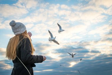 Mutlu olgun kadın, güzel uzun saçlı, beyaz örgü şapkası giyiyor mavi gökyüzüne karşı sahildeki martılar beslenir. Sonbahar ruh hali. Kopya alanı.