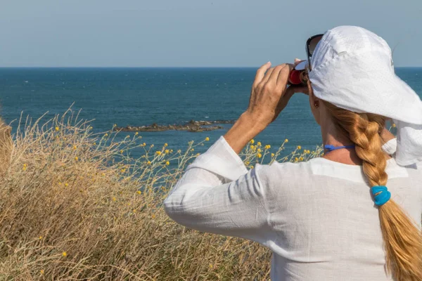 An adult woman in a white blouse and white cap, stands on the seashore and looks through binoculars.