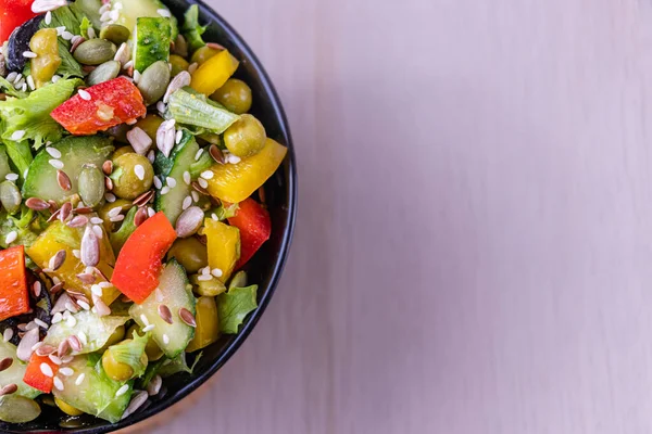 Vegetable salad in a dark bowl on a light background. Green Leaf Lettuce, cucumber, bell pepper, green peas, black and green olives, sprinkled with sunflower seeds, pumpkin seeds, sesame. Top view.
