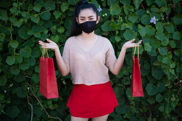 Portrait of a beautiful young Asia woman wearing a face mask with holding shopping bags at leaf wall background, Women wear pink clothes and red skirt looking at the camera