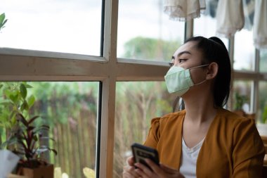 Genç Asyalı kadın oturmuş ve kafenin camından baktığı solunum hastalıklarından korunmak için tıbbi bir maske takıyor.