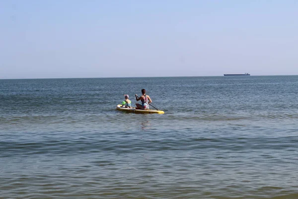 La mujer y el niño flotando sobre tabla de remo en el mar abierto donde en el horizonte un barco mercante. Deportes acuáticos, estilo de vida activo . — Foto de Stock