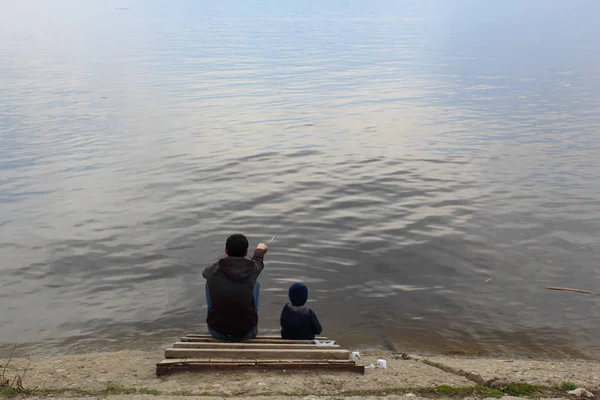 Padre e hijo se sientan en una plataforma de madera en la orilla del río y mirar a la distancia, el padre muestra algo al niño — Foto de Stock