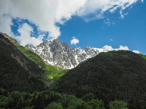 Bela paisagem montanhosa com montanhas densamente cobertas de floresta e coníferas árvores e picos rochosos parcialmente cobertos de neve no fundo do céu azul brilhante e nuvens brancas . — Fotografia de Stock