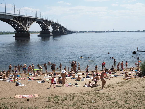 異常な暑い日に川を渡る市の橋の近くの無許可のビーチで日光浴をする人々の群衆。ロシア、サラトフ - 2019年6月 — ストック写真