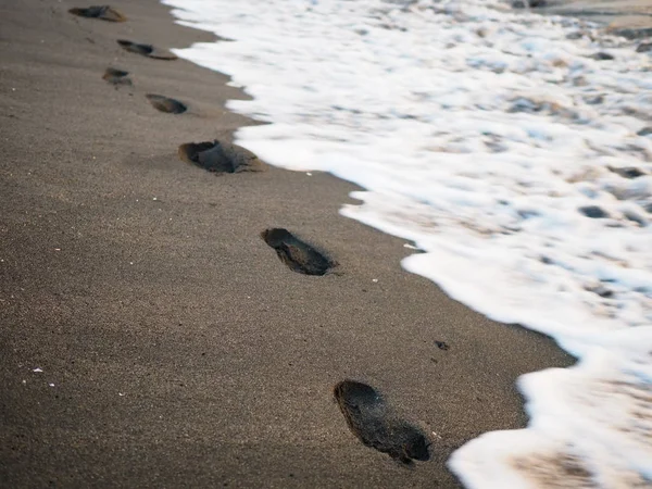 Impronte di piedi su una spiaggia di sabbia nera con sfocatura tilt-shift. Surf sullo sfondo. Concetto di solitudine, solitudine, depressione . — Foto Stock