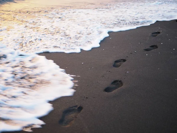 Fußabdrücke an einem schwarzen Sandstrand mit Tilt-Shift-Unschärfe. Surfen im Hintergrund. Konzept für Einsamkeit, Einsamkeit, Depression — Stockfoto