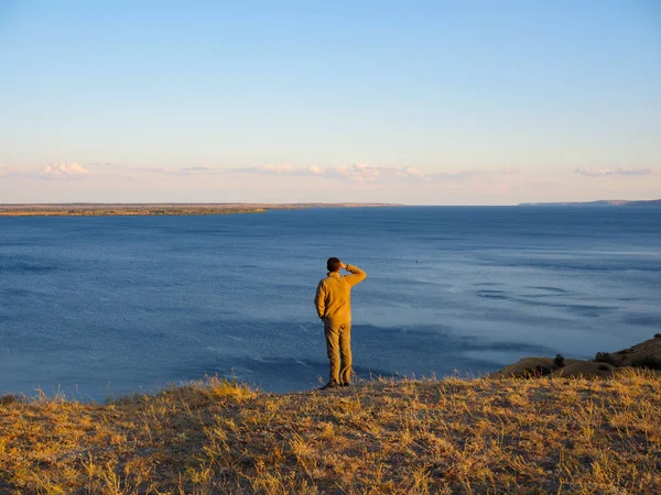 Man stands on the edge of a cliff and looks into the distance at a wide river at sunset — 스톡 사진