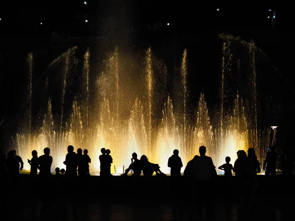 Silueta de la gente observando fuentes musicales iluminadas de colores en la noche. Luz y agua espectáculo nocturno rendimiento . — Foto de Stock