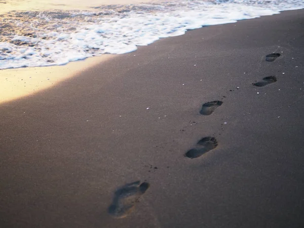 Fußabdrücke an einem schwarzen Sandstrand mit Tilt-Shift-Unschärfe. Surfen im Hintergrund. Konzept für Einsamkeit, Einsamkeit, Depression — Stockfoto