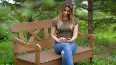 Yalnız bir bankta oturan kadın. Kızın ağaçları arasında bir bankta oturmuş smartphone kullanır. Pitoresk bir yerde bir bankta oturan kadın.