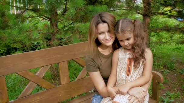 Мама сидить на лавці, вона маленька дівчинка на неї на колінах. Жінка м'яко охоплює дитини, що сидить на лавці в парку. — стокове відео