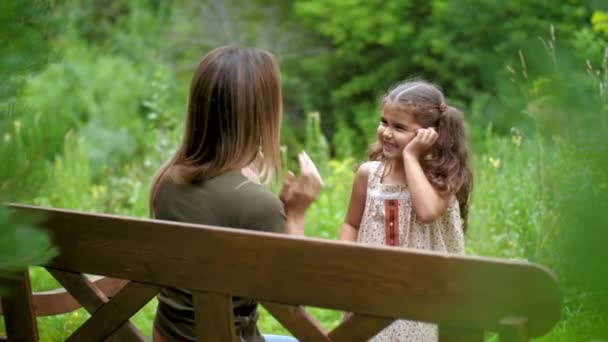 Moeder zit op een bankje onder de bomen, ze speelt pat-a-taart met het kind. Vrouw met kinderspel, zorgeloos lachen, veel plezier met het zittend op de Bank. — Stockvideo