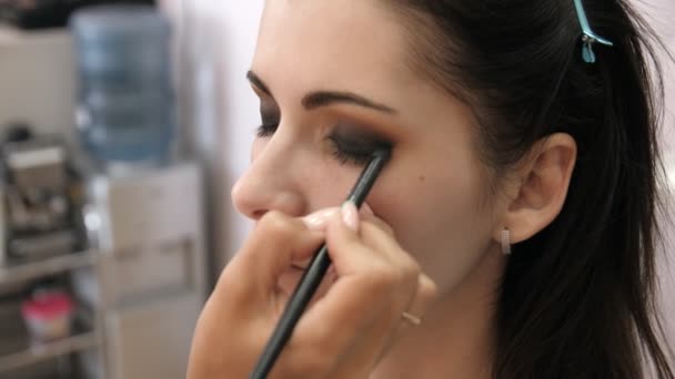Mãos maquiador aplicou maquiagem no rosto de uma jovem mulher. Menina fazendo maquiagem olho em um salão profissional. Maquiagem noturna. Olhos fumegantes. 4k — Vídeo de Stock