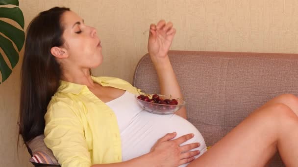 Egy fiatal terhes nő cseresznyét eszik a kanapén ülve. Gyönyörű terhes lány eszik cseresznyét egy üvegtányérról. — Stock videók