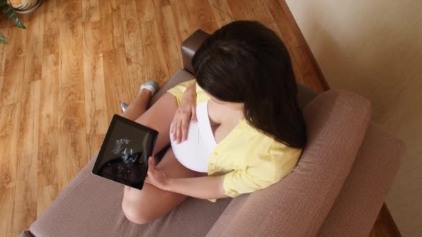 Una mujer embarazada joven está viendo un video de su ecografía en una tableta. Esperando el nacimiento, la aparición de un niño. Chica embarazada con una tableta en el sofá. Vista superior — Vídeo de stock