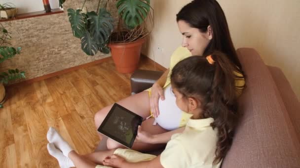 Uma jovem grávida com sua filha está assistindo a um vídeo de ultrassom em um tablet. À espera do nascimento, da aparência de uma criança. Menina grávida com um comprimido no sofá . — Vídeo de Stock