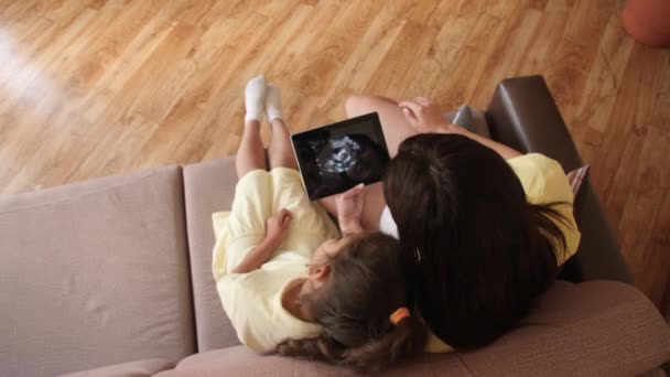 Une jeune femme enceinte avec sa fille regarde une vidéo d'échographie sur une tablette. En attendant la naissance, l'apparition d'un enfant. Fille enceinte avec une tablette sur le canapé. Vue du dessus — Video