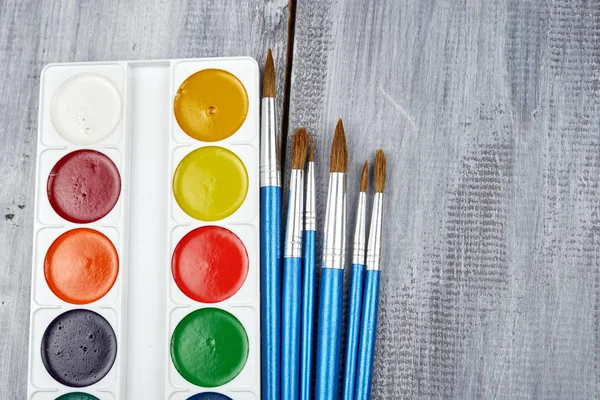 Una tavolozza di colori ad acquerello e pennelli di diverse dimensioni, distesi su uno sfondo di legno grigio — Foto Stock