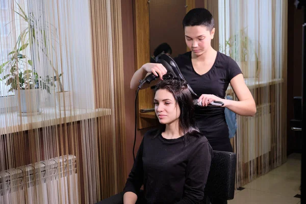 Cabeleireiro faz uma menina morena penteado em um salão de beleza com secador de cabelo . — Fotografia de Stock