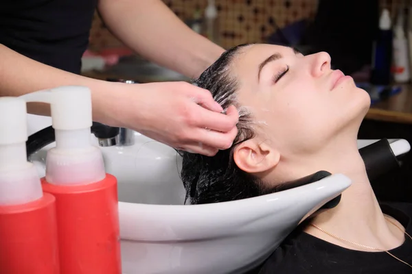 Cabeleireiro lava o cabelo com xampu para menina morena em um salão de beleza — Fotografia de Stock