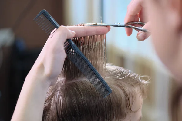 Barbeiro. Cabeleireiro faz penteado para um menino com tesoura de cabelo e pente preto . — Fotografia de Stock