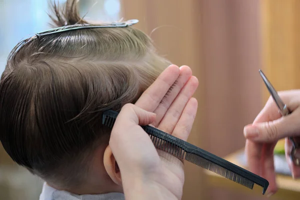 Barbeiro. Cabeleireiro faz penteado para um menino com tesoura de cabelo e pente preto . — Fotografia de Stock