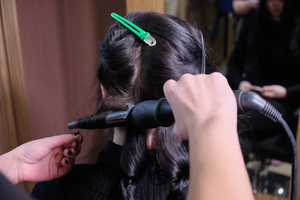 Cabeleireiro faz uma menina morena penteado em um salão de beleza com ferro Curling . — Fotografia de Stock