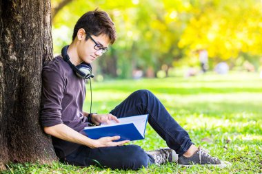 zeki ve mutlu öğrenci okuma kitap için sınav ve mutlu ruh hali ile kulaklık ile müzik dinlemek ve kampüs Üniversitesi, eğitim kavramı içinde park açık rahatlayın
