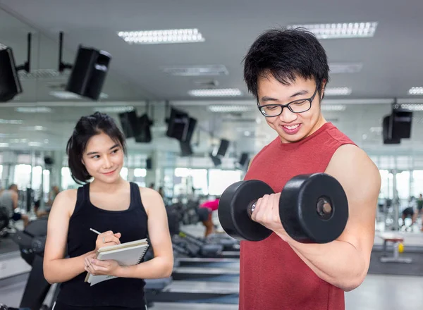 handsome young muscle man working with dumbbell with female trainer in fitness room at gym for healthy and strong with blur background of fitness equipment in concept of sport, health, and lifestyle