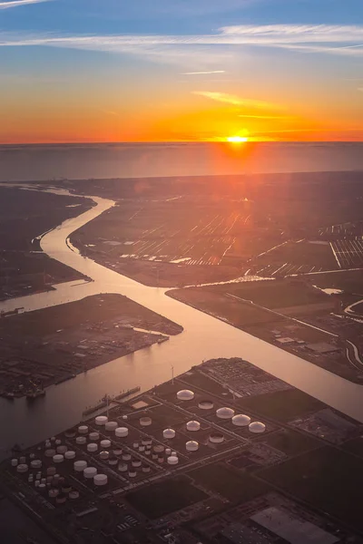 Aerial view of oil and gas tank farm in petrochemical and petroleum industrial close to river and connect to sea, ocean, for transportation at sunset time from window seat on airplane