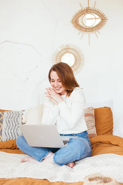 Pretty smiling woman has pleasant smile, haircut, dressed in casual domestic clothes, laugh during watching comedy, leaves comments chats online with friends on laptop computer sits in bed. — Stock Photo, Image