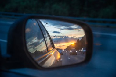 Gün batımı ve güzel gökyüzü bir arabanın aynasına yansıyor. Arabalar akşamları yüksek bir yol boyunca yol alırlar. Arabayla seyahat teması
