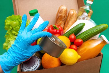 Yiyecek stoklarıyla dolu büyük bir kutu yeşillik karantina dönemi için gıda stoku. Sebze, yağ, şifalı bitkiler, izole edilmiş ekmek. Karantina, yemek teslimatı, bağış. Eldivenli bir el konserve yiyecek tutuyor..