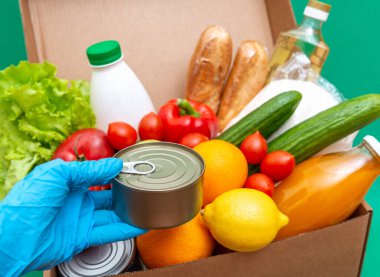 Yiyecek stoklarıyla dolu büyük bir kutu yeşillik karantina dönemi için gıda stoku. Sebze, yağ, şifalı bitkiler, izole edilmiş ekmek. Karantina, yemek teslimatı, bağış. Eldivenli bir el konserve yiyecek tutuyor..