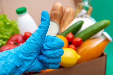 Yiyecek stoklarıyla dolu büyük bir kutu yeşillik karantina dönemi için gıda stoku. Sebze, yağ, şifalı bitkiler, izole edilmiş ekmek. Karantina, yemek teslimatı, bağış. Eldiven eldiveni başparmağı gösteriyor..