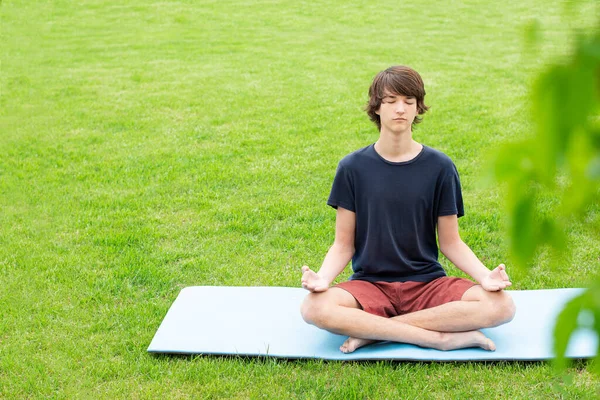 A young man with long hair does yoga outdoors. A teenager sits in a lotus position on the green grass. Copy space. Place for text.