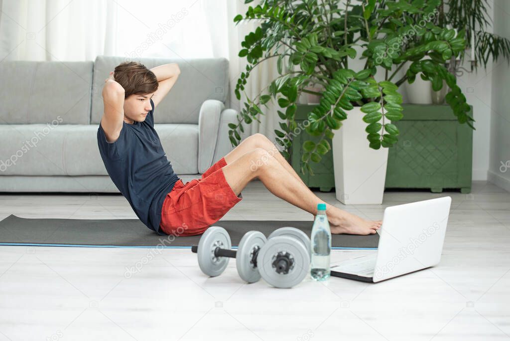 Handsome young man in a dark t-shirt and red shorts does sports at home online. Teenager is training in the room