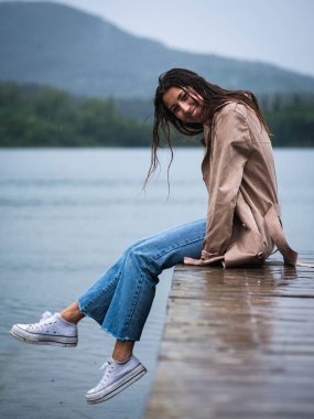 Genç bir kadın güzel bir gölde, yağmurda, ahşap bir iskelede oturuyor.