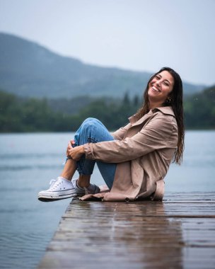 Genç bir kadın güzel bir gölde, yağmurda, ahşap bir iskelede oturuyor.