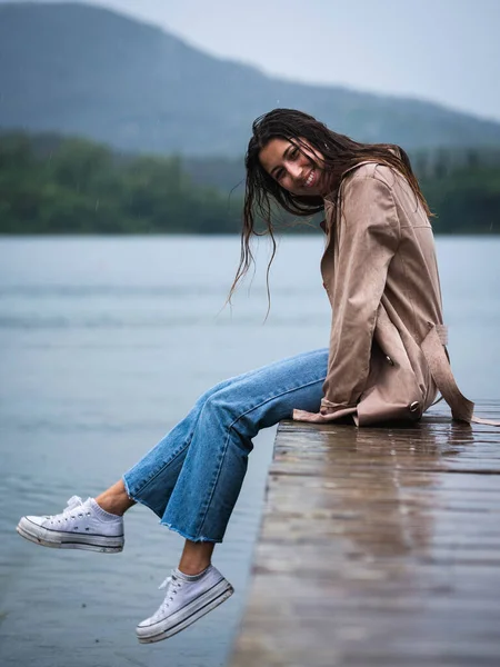 Genç bir kadın güzel bir gölde, yağmurda, ahşap bir iskelede oturuyor.