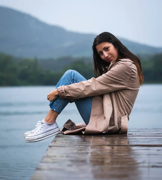 Genç bir kadın güzel bir gölde, yağmurda, ahşap bir iskelede oturuyor.